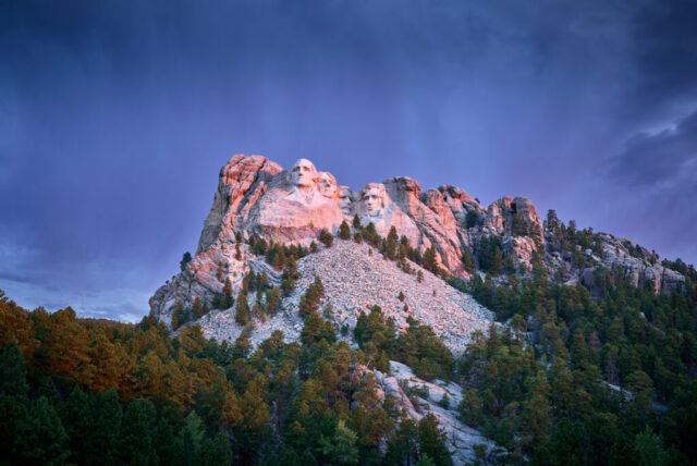 Panoramic landscape photograph of Mount Rushmore in South Dakota at sunrise showing warm light on the carved faces and contrasting blue clouds in the sky