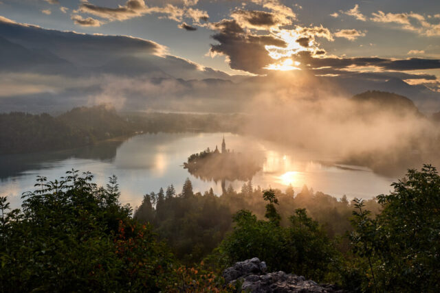 Sunrise landscape photograph of Lake Bled in Slovenia showing mist over the lake, reflections of light on the water, and the island with its church viewed from a hilltop viewpoint