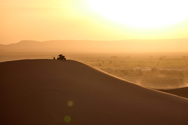 Golden hour backlit image of the Merzouga dunes in the Sahara Desert, Morocco, showing a person sitting on the dune crest and a buggy below