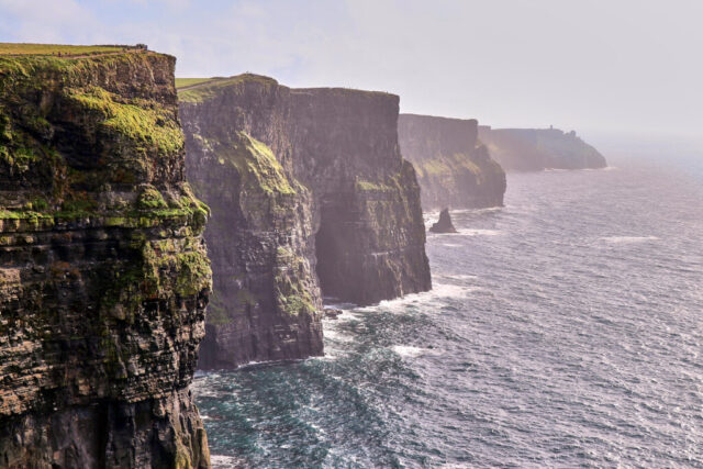 Photograph of the Cliffs of Moher in Ireland showing green cliffs under heavy clouds with limited sunlight over the Atlantic Ocean