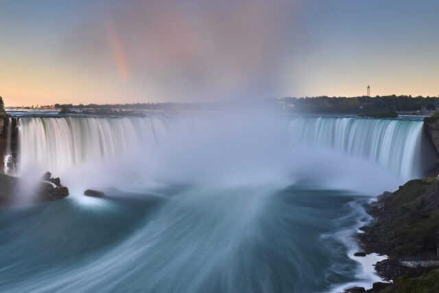 Long exposure image of Niagara Falls at sunset showing flowing water, mist, and a rainbow in the sky